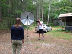 Tomy O'Brien, standing, focuses his camera and flash umbrella at Caragh O'Brien, with car, forest, and cabin in background.